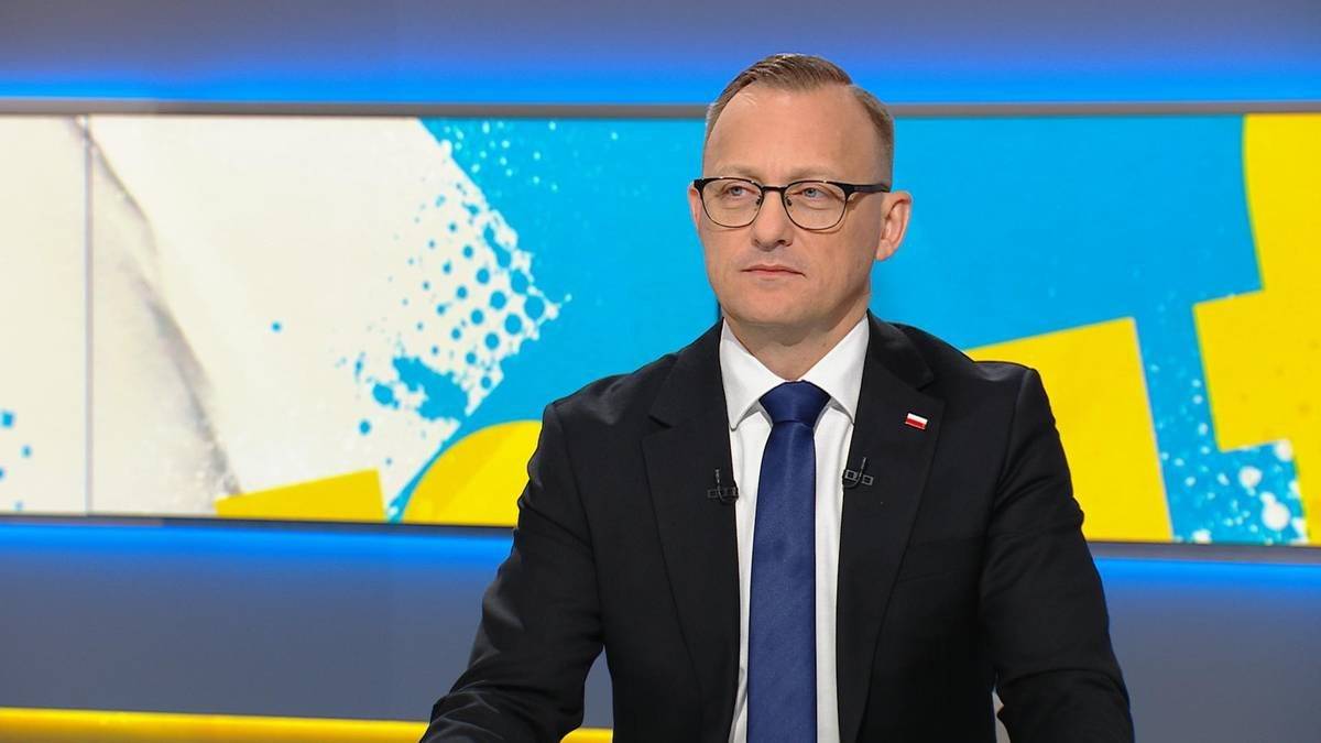 Mężczyzna w okularach, czarnym garniturze i granatowym krawacie siedzi i patrzy w bok. W tle znajduje się jasny ekran z abstrakcyjnym wzorem w kolorach niebieskim, białym i żółtym.