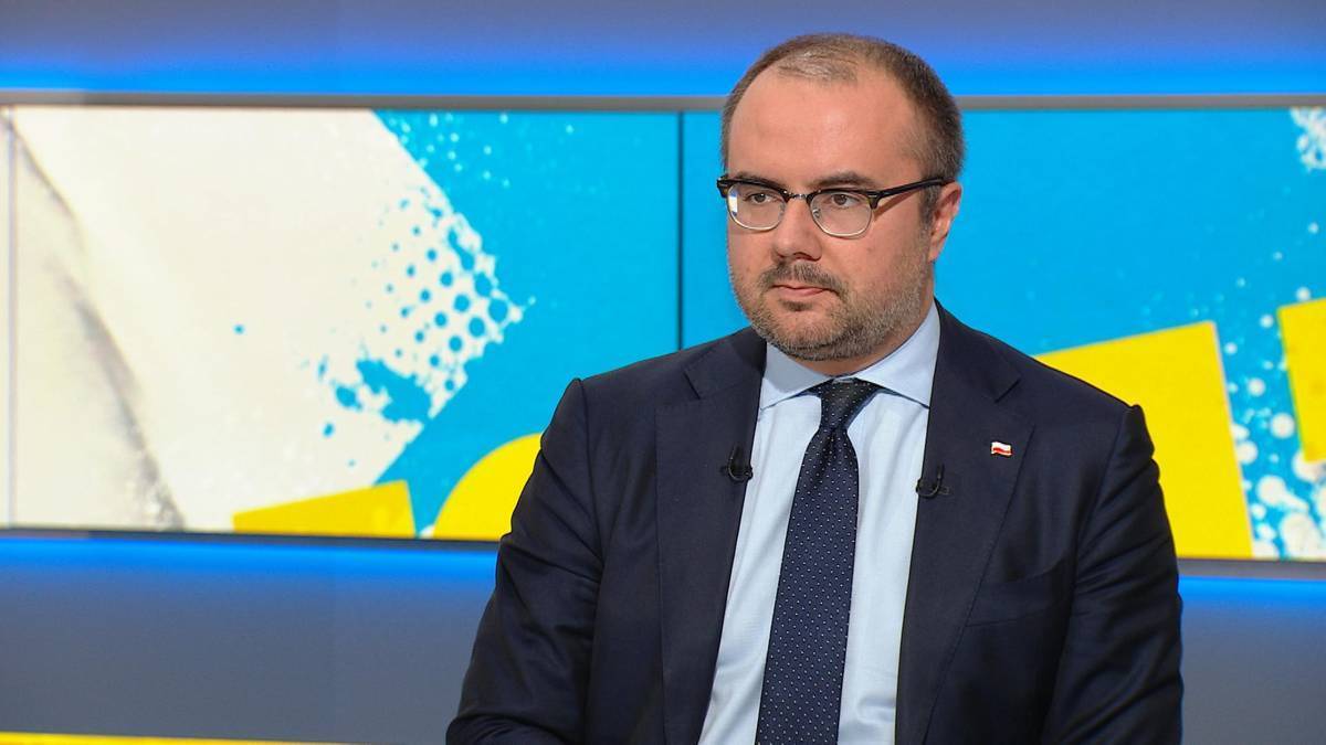 Mężczyzna w okularach i garniturze siedzi przed ekranem telewizyjnym.