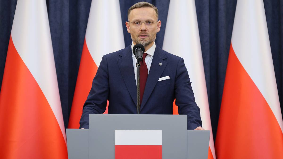 Zbigniew Bogucki przemawia za m&oacute;wnicą przed flagami Polski.