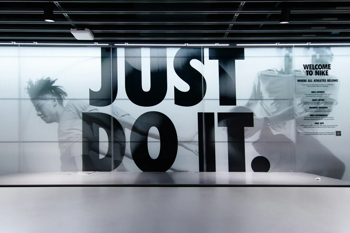 Wnętrze sklepu Nike z wielkim hasłem „Just Do It” na ścianie i minimalistycznym stylem. Zniżka wartości akcji Nike kontrastuje z mocnym wizerunkiem przedsiębiorstwa, który przez lata kształtował jej ogólnoświatową pozycję.