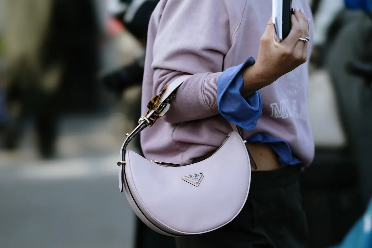 Osoba w stroju streetwearowym trzyma jasnofioletową torebkę firmy Prada na ramieniu, uwieczniona na zdjęciu podczas pokazu mody na ulicy. Gdzie można zaoferować luksusową odzież online?