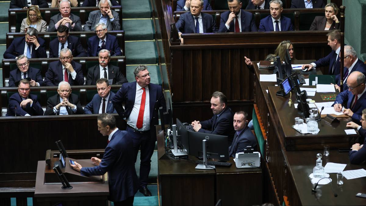 Grupa polityk&oacute;w siedzących w ławach sejmowych, jeden z nich stoi i przemawia.