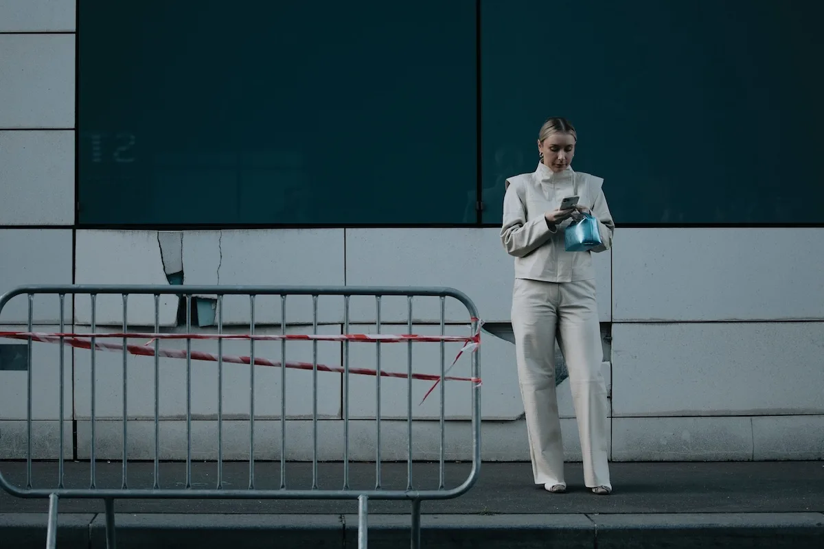 Kobieta w jasnym ubraniu stoi obok nowoczesnego budynku i korzysta z telefonu. Obraz podkreśla powszechne używanie technologii i kontekst cyfrowych informacji, które mogą być narażone na przecieki.