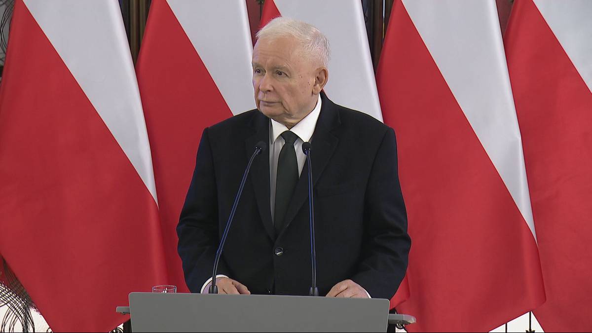 Jarosław Kaczyński przemawia za m&oacute;wnicą, na tle flag Polski.