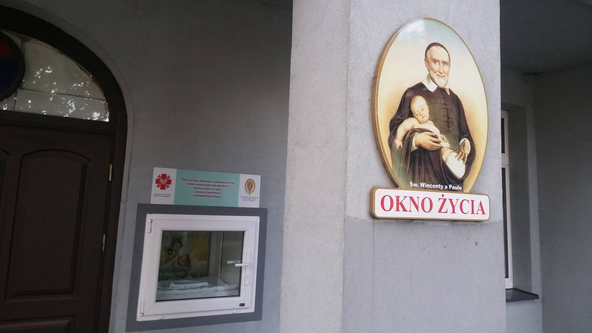Owalny medalion z postacią świętego trzymającego dziecko, pod nim napis "Okno życia". Obok znajduje się białe, prostokątne okno życia.