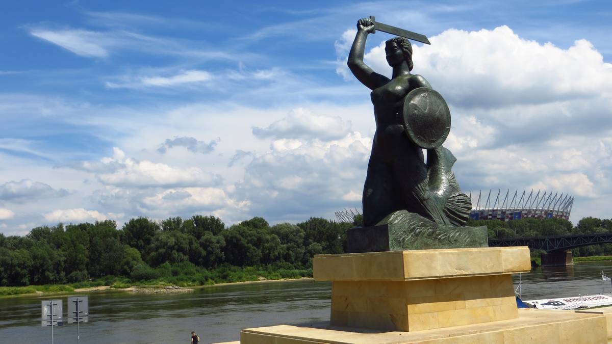 Pomnik Warszawskiej Syrenki z mieczem i tarczą na cokole, na tle rzeki, drzew i nieba z chmurami.