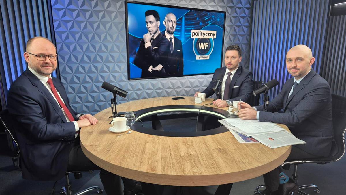 Trzech mężczyzn w garniturach siedzi przy okrągłym stole w studiu telewizyjnym. Na ścianie za nimi wyświetlany jest ekran z logo programu "Polityczny WF".