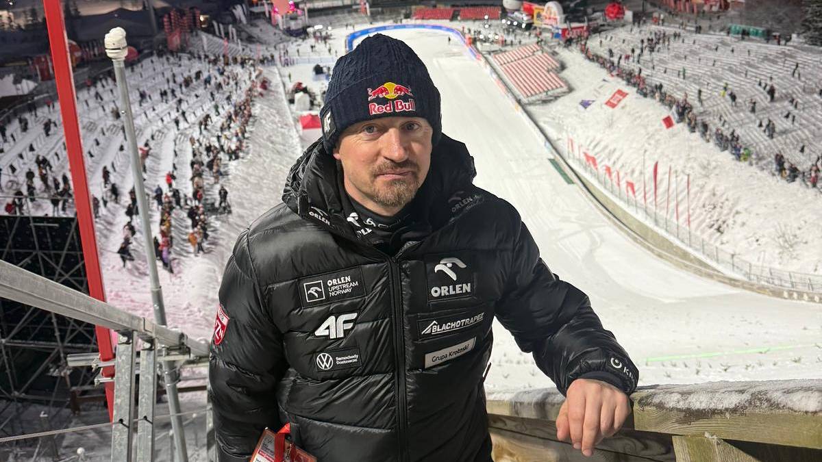 Portret Adama Małysza w kurtce zimowej, stojącego na tle skoczni narciarskiej nocą.