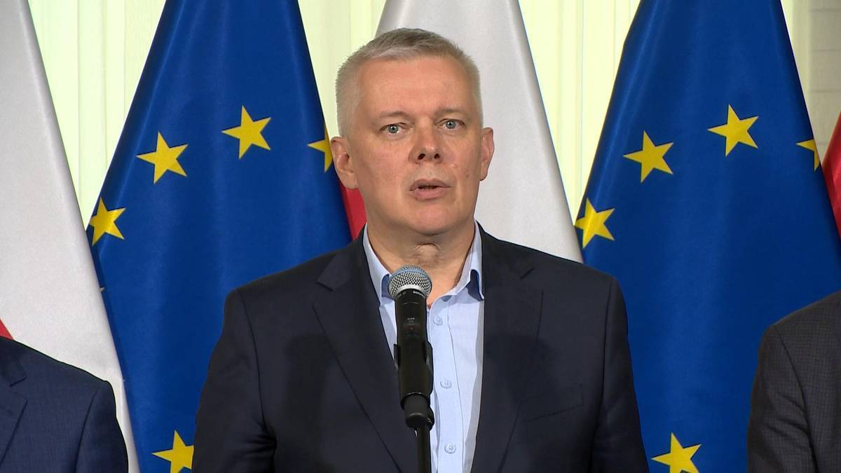 Mężczyzna w garniturze przemawia do mikrofonu na tle flag Unii Europejskiej.