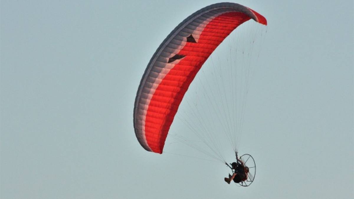 Paralotnia z czerwonym i szarym skrzydłem lecąca na tle błękitnego nieba, z widocznym pilotem pod skrzydłem.