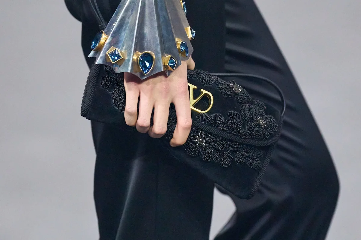 Zbliżenie na czarną, zdobioną kopertówkę Valentino trzymaną w dłoni modelki, z widocznym złotym logo oraz bogato dekorowanym mankietem z niebieskimi kamieniami. Elegancki detal podkreśla luksusowy charakter marki, która jednocześnie mierzyła się z nadzorem sądowym związanym z nieprawidłowościami w łańcuchu dostaw. Kontrast między estetyką produktu a kulisami produkcji przypomina o znaczeniu transparentności w branży premium.