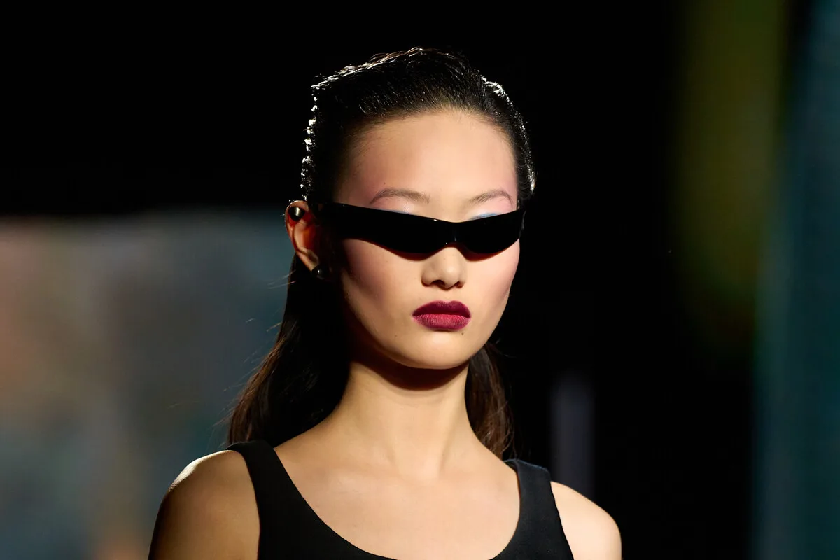 Modelka na pokazie prezentuje prosty, lecz efektowny make-up – z intensywnie zaakcentowanymi wargami i delikatnym wykończeniem cery, dopełniony futurystycznymi okularami. Ten wygląd przywodzi na myśl stylistykę awatarów w Roblox, gdzie make-up często zestawia minimalizm z charakterystycznym detalem. Stylizacja ilustruje, w jaki sposób aktualna moda i uroda inspirują wirtualną indywidualizację postaci.