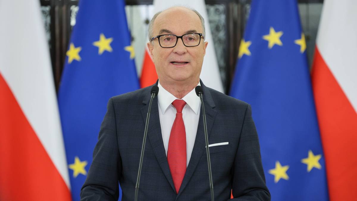 Mężczyzna w okularach i czerwonym krawacie przemawia za m&oacute;wnicą, na tle flag Polski i Unii Europejskiej.