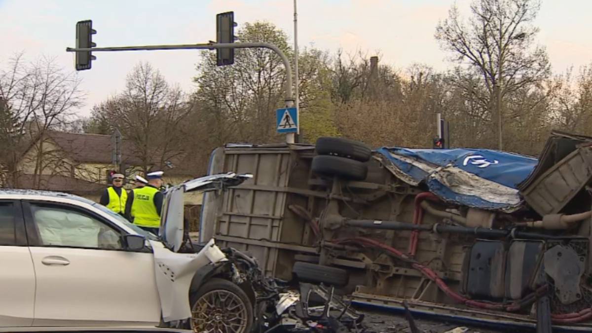 Wypadek drogowy ze skutkiem przewrócenia się autobusu na bok, obok uszkodzony samochód osobowy i stojący policjanci.