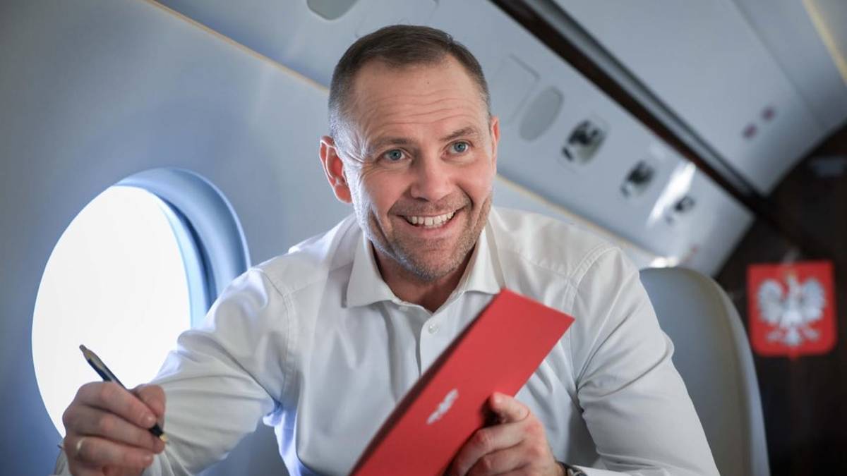 Mężczyzna w białej koszuli siedzi w samolocie, trzymając w rękach tablet i długopis.