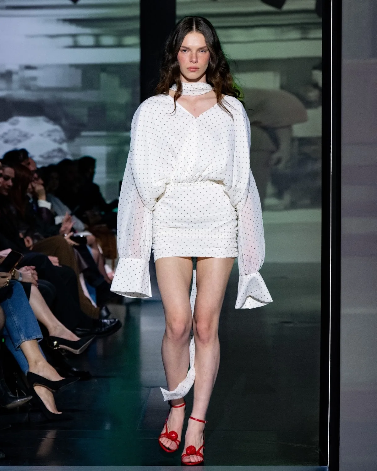 Modelka idzie wybiegiem w białej, lekkiej sukience mini w delikatne kropki, z bufiastymi rękawami i subtelnym wiązaniem pod szyją. Stylizację przełamują czerwone sandały na obcasie, które dodają kontrastu i podkreślają kobiecy, świeży charakter outfitu.