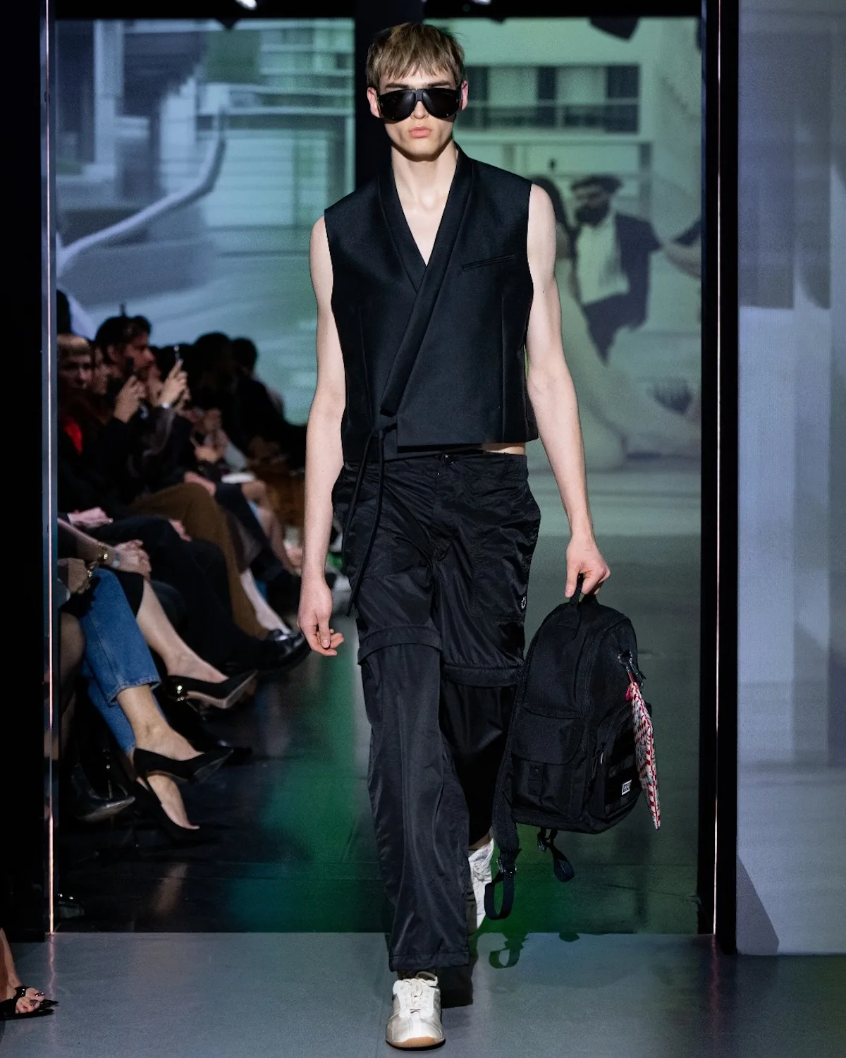 Model prezentuje nowoczesną, minimalistyczną stylizację w czerni – bezrękawnik o kroju inspirowanym marynarką zestawiono z luźnymi spodniami i sportowymi butami. Całość uzupełniają duże okulary przeciwsłoneczne oraz plecak, nadając lookowi miejski, funkcjonalny charakter.