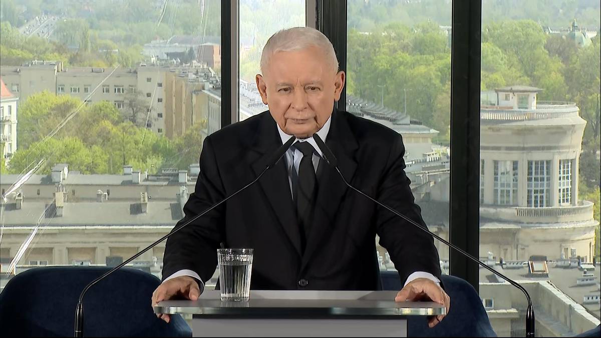 Jarosław Kaczyński, lider PiS, ubrany w czarny garnitur i białą koszulę z czarnym krawatem, stoi przy m&oacute;wnicy z mikrofonami i szklanką wody na tle panoramicznego okna z widokiem na miasto.