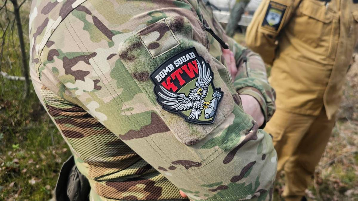 Zbliżenie na rękaw munduru wojskowego w kamuflażu z naszywką "BOMB SQUAD KTW" przedstawiającą orła z rozpostartymi skrzydłami.