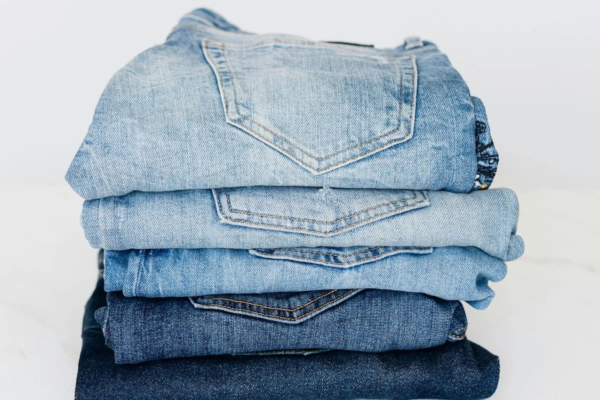 Stos dżinsów w różnych odcieniach błękitu ułożonych jeden na drugim, widok na tylne kieszenie spodni denimowych.