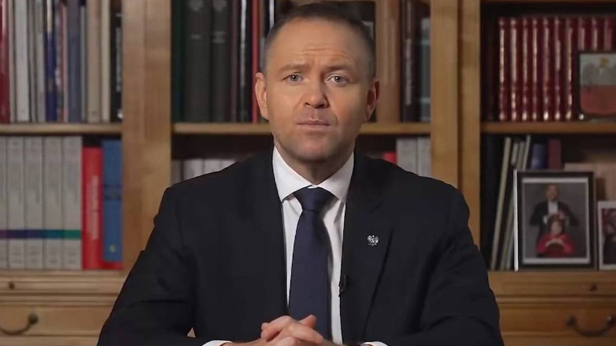 Karol Nawrocki siedzi przy biurku na tle regałów z książkami.