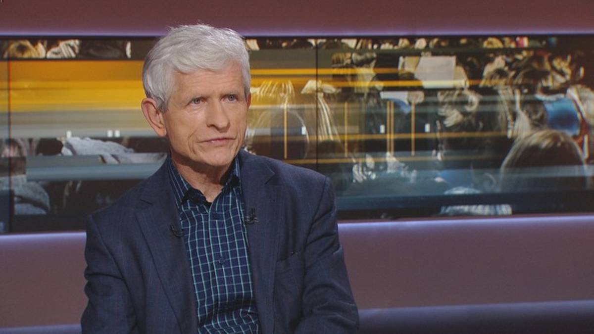 Mężczyzna w średnim wieku z siwymi włosami, w granatowej marynarce i pasiastym niebiesko-białym swetrze, siedzi na tle ekranu z ruchomymi obrazami.