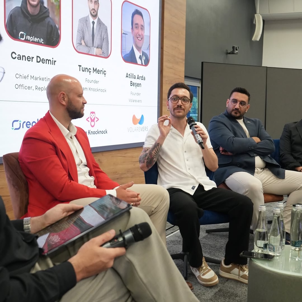 Panel dyskusyjny startupu Replenit – kilku mężczyzn siedzi na scenie, jeden z nich prowadzi wypowiedź do mikrofonu, a w tle widoczna jest tablica z nazwiskami oraz logo firmy.