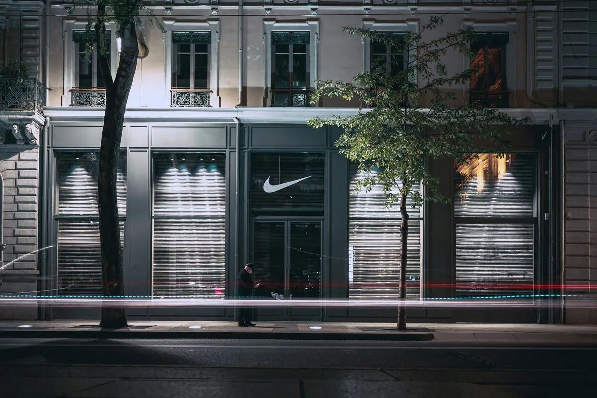 Zamknięty sklep Nike z zasuniętymi roletami i logo na witrynie w miejskiej przestrzeni nocą. Zniżka wartości akcji Nike odzwierciedla trudniejszy czas dla przedsiębiorstwa i zmieniającą się sytuację rynkową.