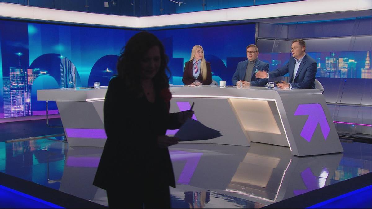 Trzy osoby siedzą za stołem w studiu telewizyjnym, a jedna osoba stoi przed stołem, trzymając dokument.