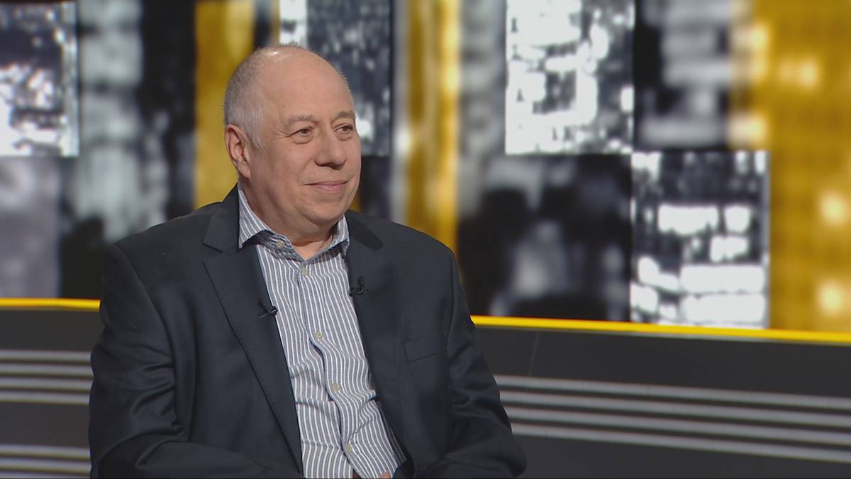 Mężczyzna w garniturze siedzi na tle nowoczesnego studia telewizyjnego.