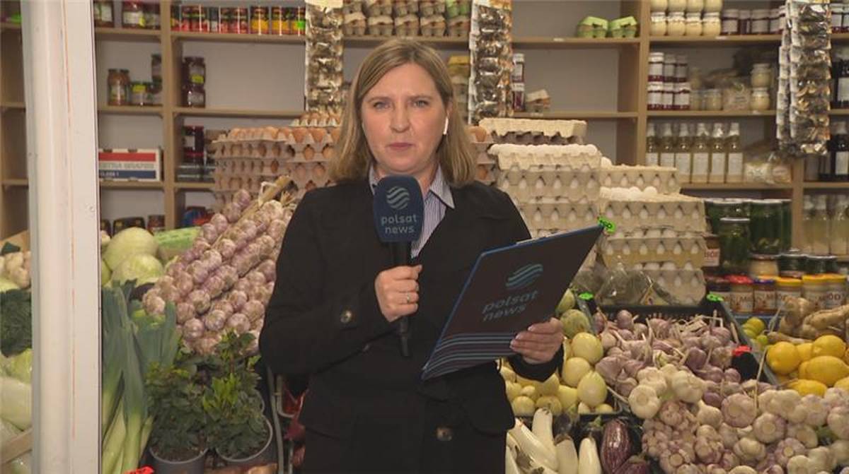 Kobieta w czarnym stroju trzymająca mikrofon Polsat News i tablet, stoi przed stoiskiem z produktami spożywczymi.