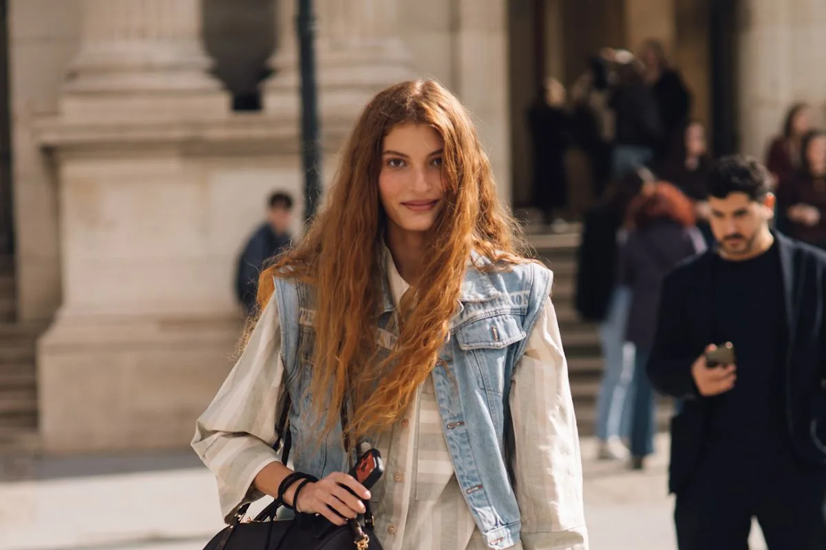 Fotografia prezentuje stylową niewiastę z długimi, rudymi włosami, stojącą na placu i uśmiechającą się w stronę obiektywu. Ubrana jest w beżową bluzkę w prążki, dżinsową kurtkę bez rękawów, luźne spodnie jeansowe typu baggie i czarno-białe sportowe buty na platformie. W dłoni dzierży czarną torebkę z widocznym wisiorkiem. Ten nowoczesny i trendowy wygląd doskonale koresponduje z aktualnymi tendencjami w modzie. Jeżeli w Twojej garderobie znajdują się podobne unikaty, sprawdź, gdzie zaoferować ubrania w sieci.