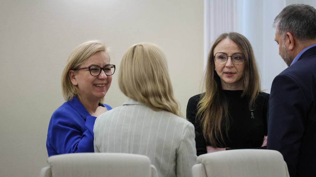 Cztery osoby stojące w pomieszczeniu, dwie kobiety rozmawiają ze sobą, pozostałe osoby patrzą w ich stronę.