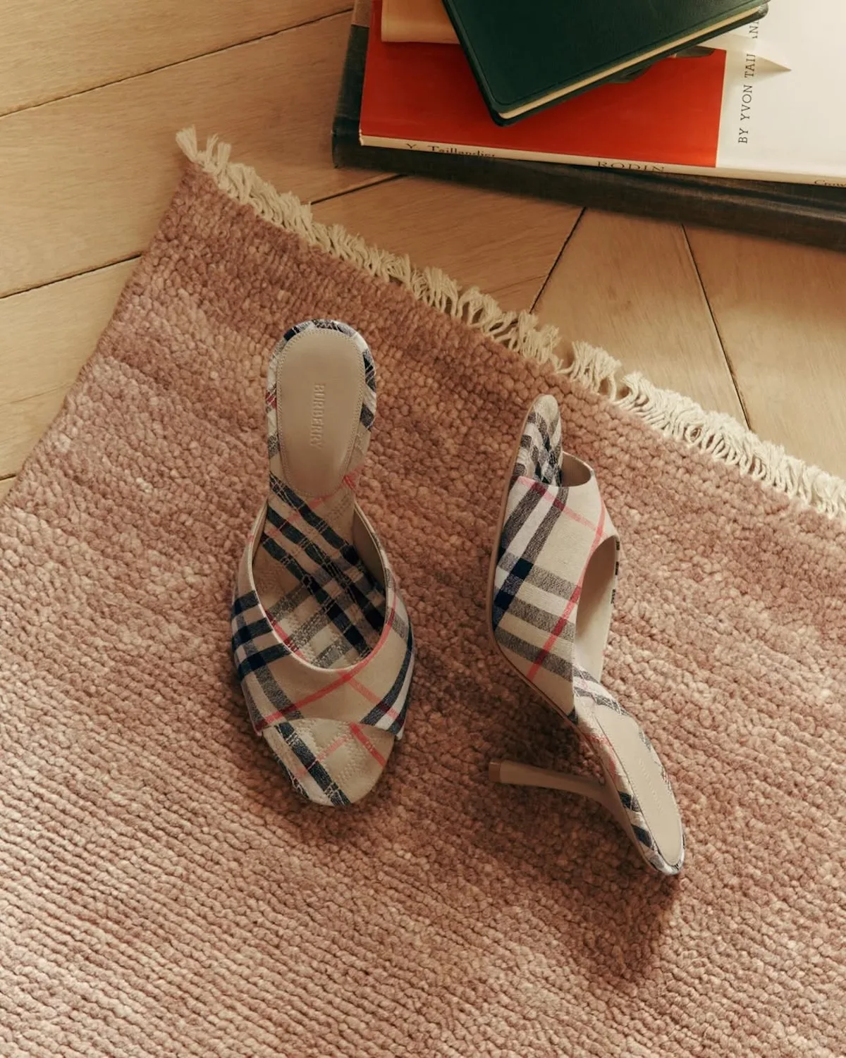 Buty na obcasie Burberry w charakterystyczną kratę leżą na jasnoróżowym dywanie obok książek. Zdjęcie podkreśla znaczenie rozpoznawalnych wzorów i akcesoriów w ofercie brytyjskiego domu mody.