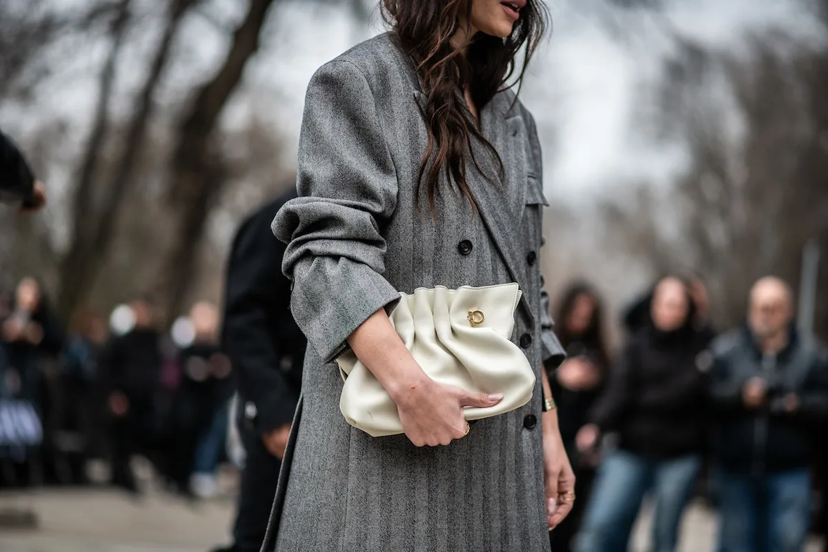 Kobieta w eleganckim szarym płaszczu z jasną torebką podczas wydarzenia modowego w Europie. Zdjęcie nawiązuje do zmian w branży fashion, gdzie nowe regulacje UE dotyczące zakazu produktów z pracy przymusowej zwiększają znaczenie transparentnych łańcuchów dostaw. Marki modowe muszą dziś nie tylko projektować, ale też udowadniać etyczne pochodzenie swoich produktów.