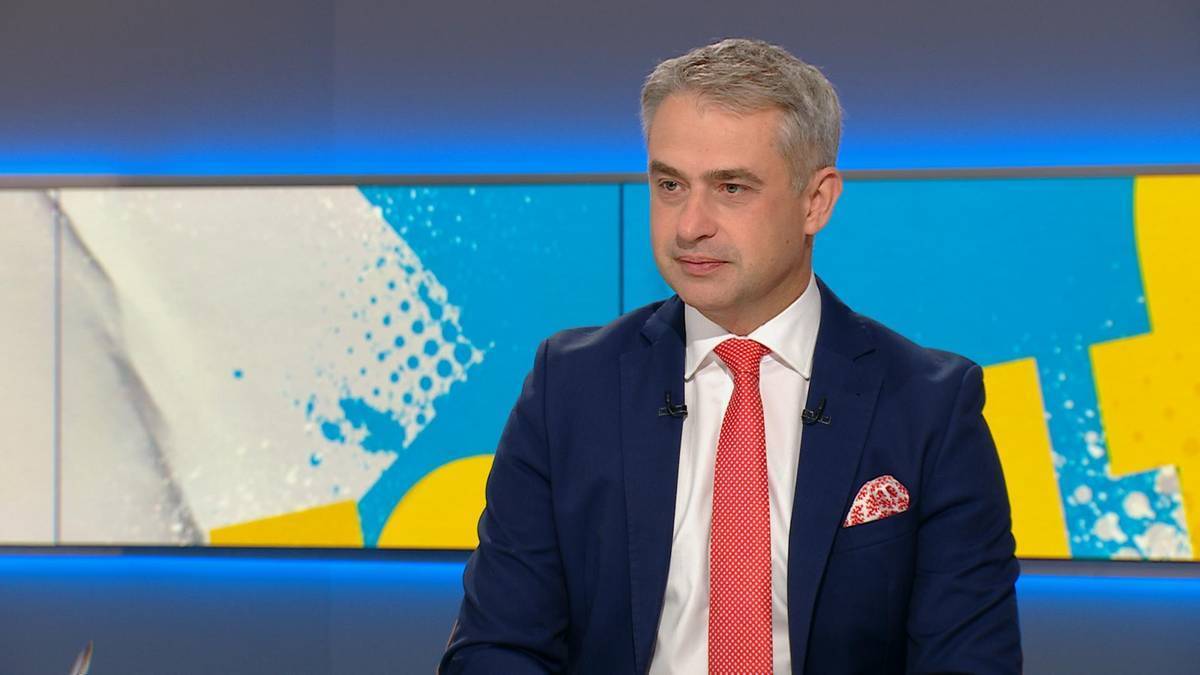 Mężczyzna w granatowym garniturze i czerwonym krawacie siedzi w studiu telewizyjnym.