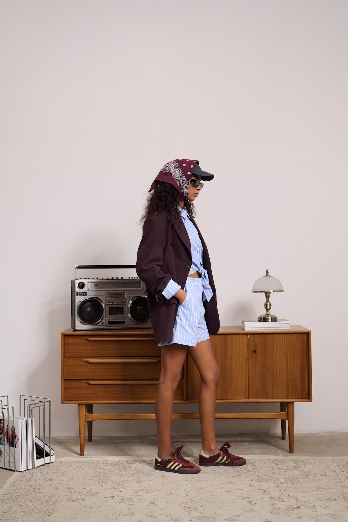 Modelka w bordowej marynarce MISAGA, błękitnym komplecie w paski i sneakersach w stylu retro na tle drewnianej komody.