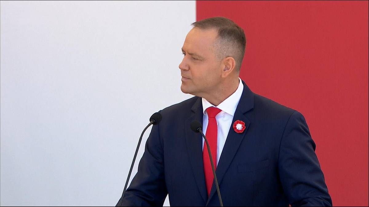 Mężczyzna w garniturze i czerwonym krawacie przemawia za m&oacute;wnicą, na tle biało-czerwonych pas&oacute;w.