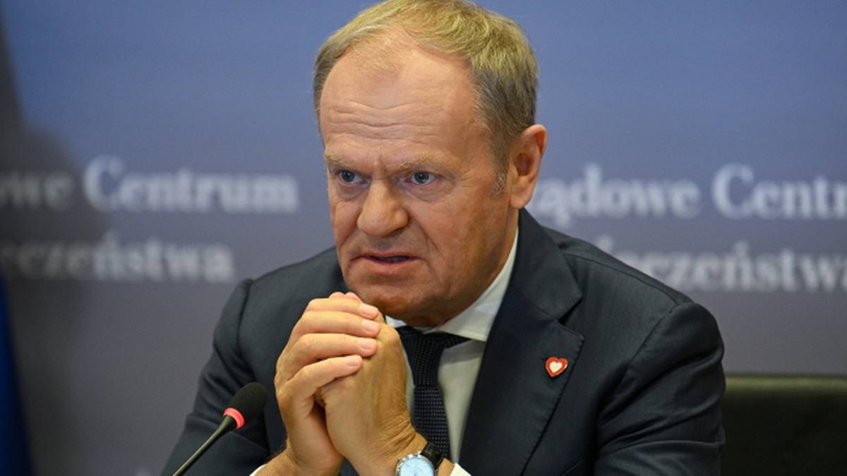 Donald Tusk trzyma złożone dłonie przed sobą, siedząc przy stole.