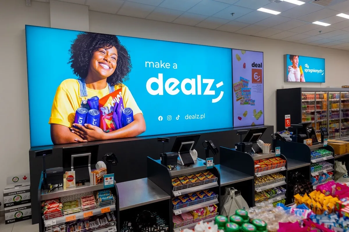 Wnętrze sklepu Dealz z kasami, regałami ze słodyczami i dużą reklamą marki na ścianie. Sprzedaż Dealz Poland za symboliczną kwotę 1 zł to jeden z najbardziej komentowanych ruchów na rynku handlu detalicznego.