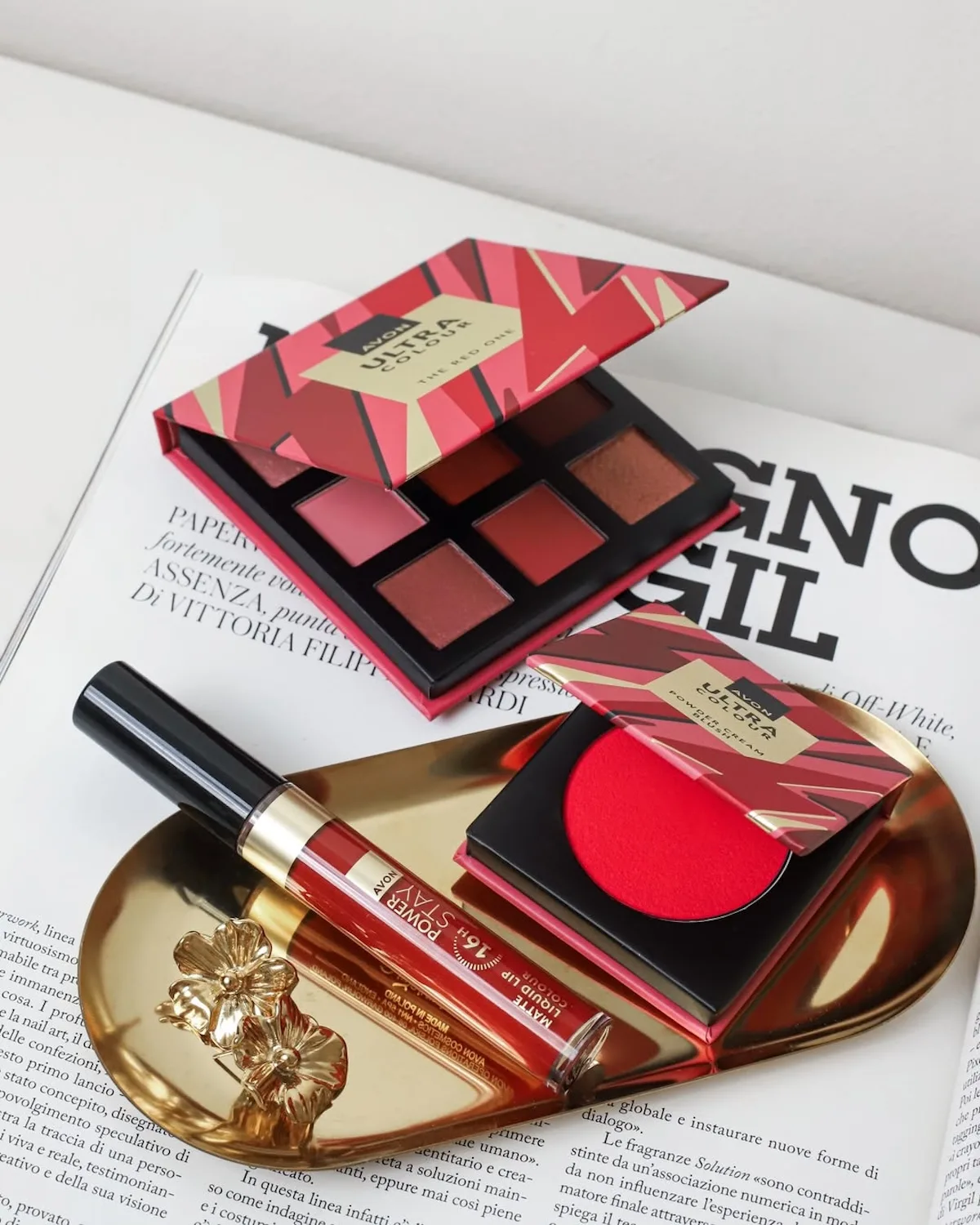 Na zdjęciu widać kosmetyki Avon ułożone na złotej tacce i otwartym magazynie. W kadrze znajdują się m.in. palety do makijażu, róż oraz matowa pomadka w czerwonym odcieniu.