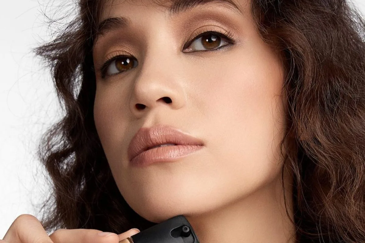 Modelka prezentuje naturalny makijaż i promienną cerę, a w kadrze widoczne jest opakowanie kosmetyku do makijażu. Fotografia ilustruje artykuł o MAC Cosmetics, pozwie zbiorowym w USA, technologii virtual try-on oraz zarzutach dotyczących skanowania twarzy i danych biometrycznych.