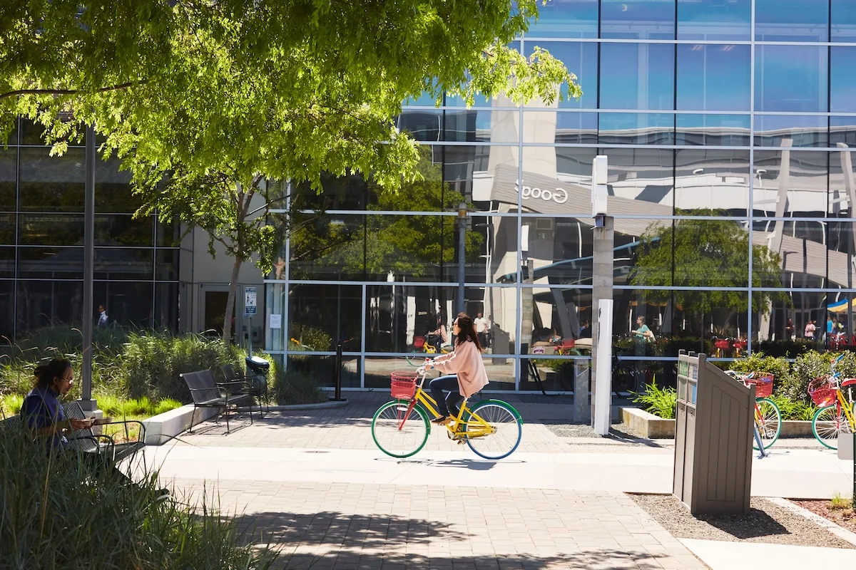 Kobieta na kolorowym rowerze przed nowoczesnym biurowcem Google. W tle widać zieleń, stojaki rowerowe i odbicia budynków w szklanej fasadzie.
