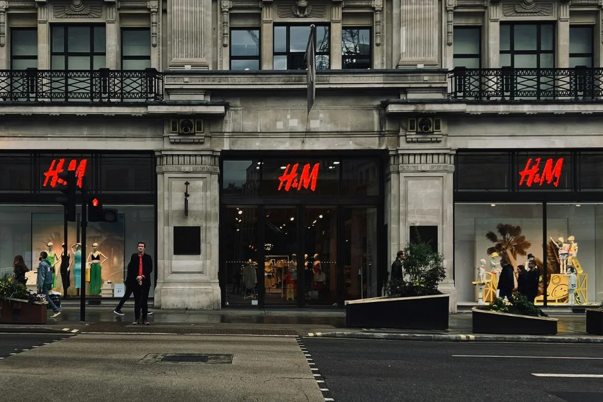 Na zdjęciu widać fasadę sklepu H&amp;M przy miejskiej ulicy, z czerwonym logo marki nad wejściem i w witrynach. Przed salonem przechodzą piesi, a elegancka kamienica podkreśla stacjonarny charakter lokalizacji. Fotografia nawiązuje do tematu zmian w sieci sprzedaży H&amp;M i selektywnego podejścia do utrzymywania sklepów w najlepszych adresach.