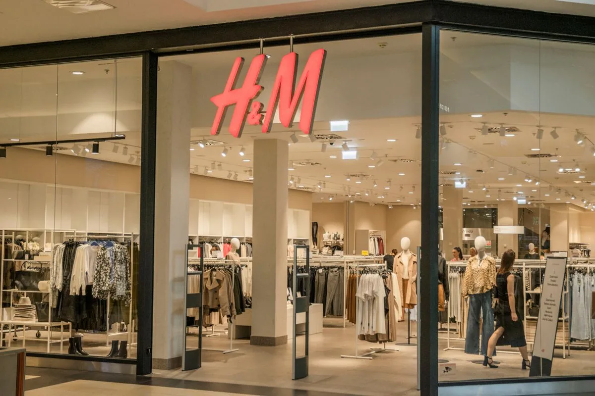 Na zdjęciu widać wejście do sklepu stacjonarnego H&M w centrum handlowym, z czerwonym logo marki nad witryną. W środku widoczne są wieszaki z ubraniami, manekiny i jasne, nowoczesne wnętrze sklepu.