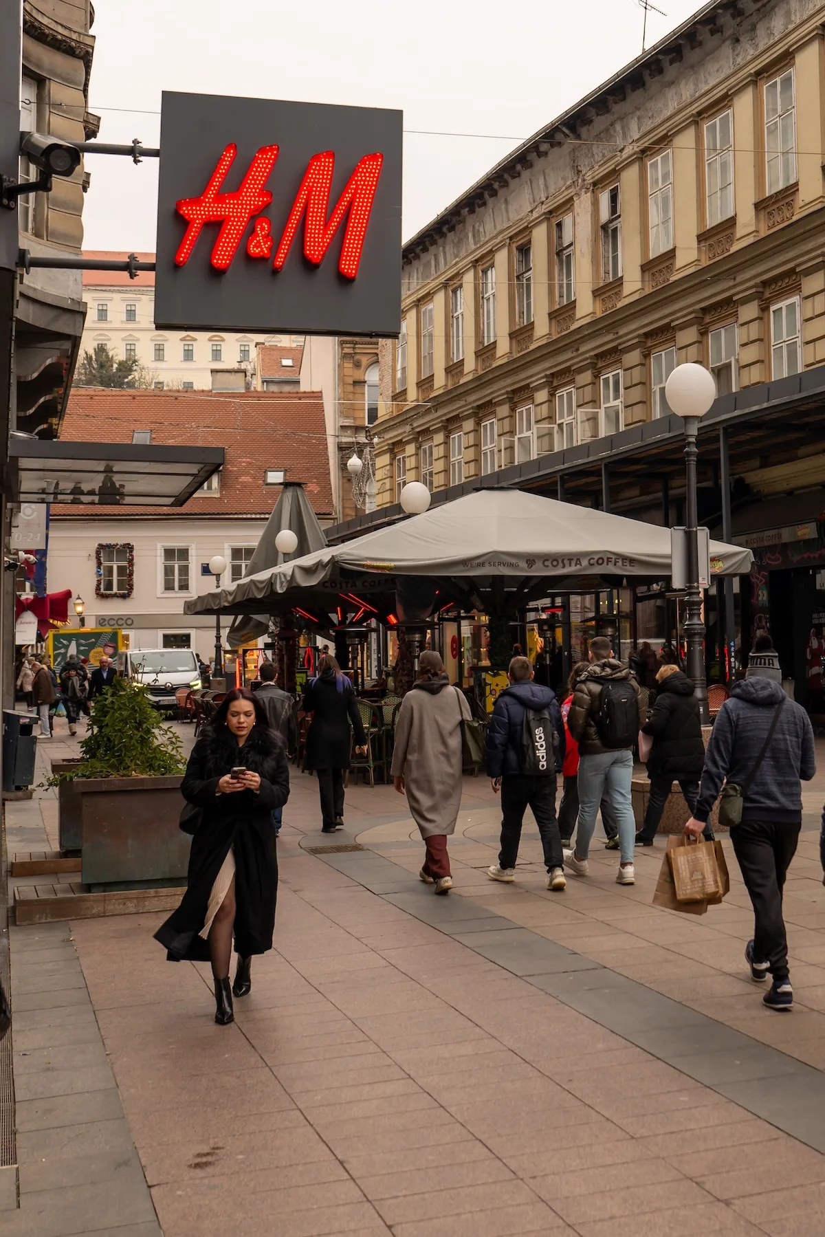 Na zdjęciu widać duży, czerwony szyld H&M nad ruchliwą ulicą handlową z przechodniami i ogródkami kawiarni. Fotografia nawiązuje do tematu sklepów stacjonarnych marki, których liczba ma być stopniowo ograniczana w ramach zmiany strategii sprzedaży.