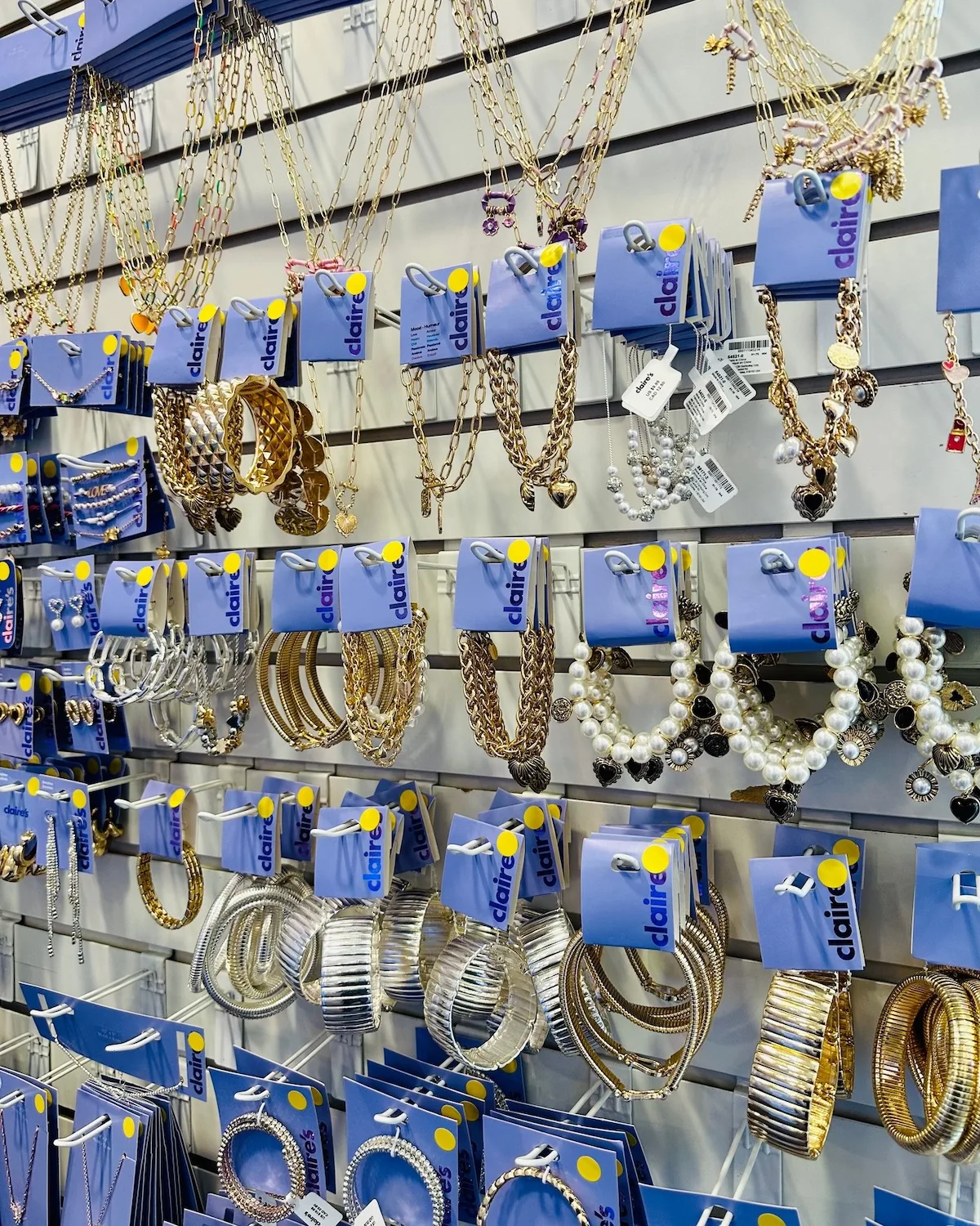 Ekspozycja biżuterii i akcesoriów w sklepie Claire’s. Na ścianie widać naszyjniki, bransoletki i ozdoby na charakterystycznych niebiesko-fioletowych kartonikach marki.