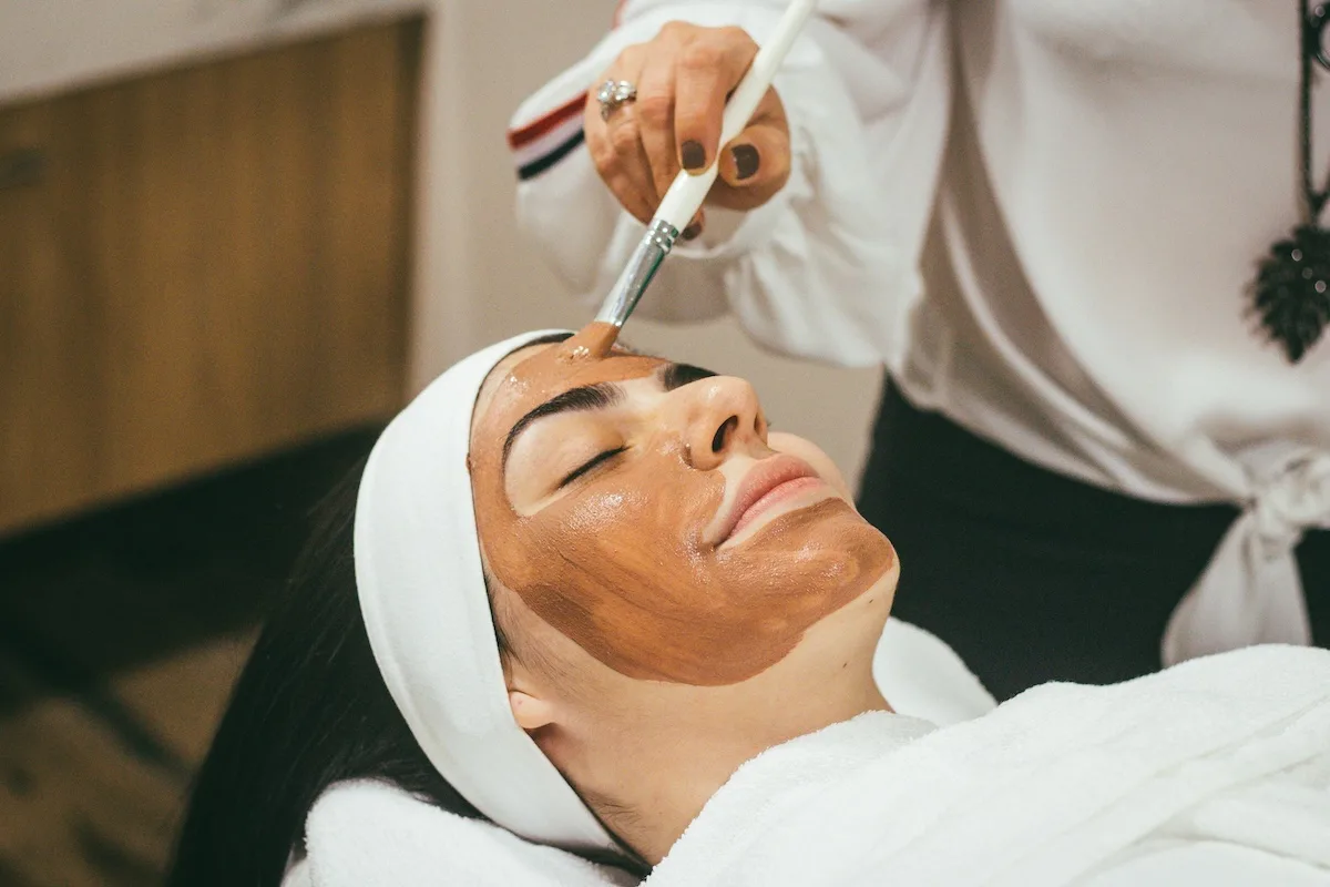 Kosmetyczka nakłada pędzlem maseczkę na twarz klientki podczas zabiegu pielęgnacyjnego w salonie beauty. Zdjęcie pokazuje profesjonalną usługę kosmetyczną i dobrze ilustruje temat rezerwacji wizyt w branży urody.