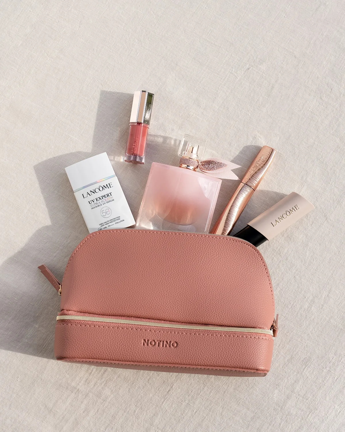 Różowa kosmetyczka Notino leży na jasnym materiale, a z jej wnętrza wystają kosmetyki i perfumy Lancôme. Zdjęcie podkreśla połączenie pielęgnacji, makijażu i zakupów beauty w jednym estetycznym ujęciu.