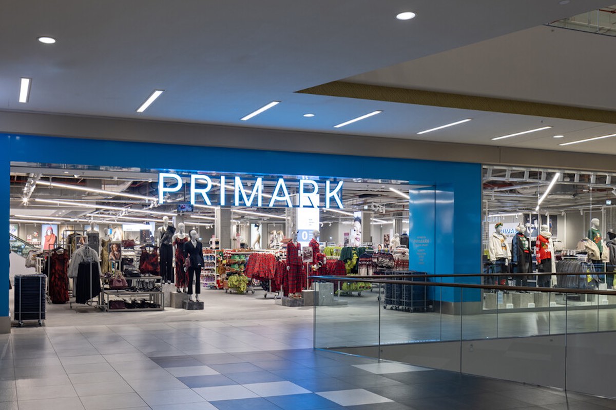 Wejście do sklepu Primark w galerii handlowej, z dużym podświetlonym logo „PRIMARK” nad szerokim otwartym przejściem. Wewnątrz widoczne są manekiny ubrane w różne stylizacje oraz regały z ubraniami i akcesoriami.